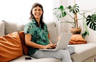 Eine Frau sitzt auf einer hellen Couch mit orangefarbenen Kissen, Laptop auf dem Schoß und Smartphone daneben.