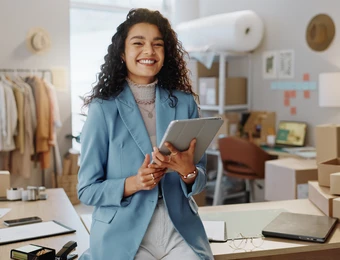 Eine Frau mit lockigen Haaren in blauem Blazer hält Tablet, lächelt; Hintergrund: Kartons, Laptop, Kleiderständer und Lampen.