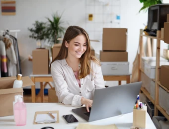 Frau sitzt am Laptop an einem Schreibtisch, umgeben von Kartons, Regalen und Bürobedarf.