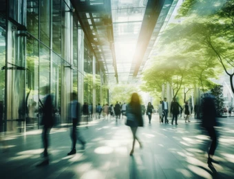 Gläserne Gebäudefassade mit belebter, unscharf wirkender Fußgängerzone; Menschen gehen, Sonnenlicht strahlt durch Glasdach.