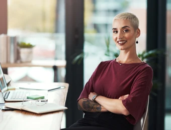 Person mit kurzem blondem Haar, maroonfarbenes Oberteil, sitzt am Schreibtisch mit Laptop, Arme verschränkt, lächelnd.