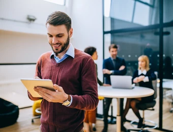Mann in rotem Pullover hält Tablet; im Hintergrund mehrere Personen um einen runden Tisch mit Laptop in einem modernen Büro.