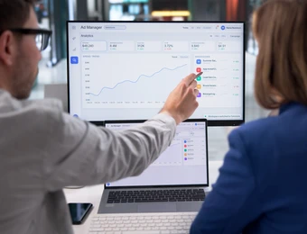 Zwei Personen schauen auf einen Monitor mit Diagrammen; eine zeigt auf die Grafik.