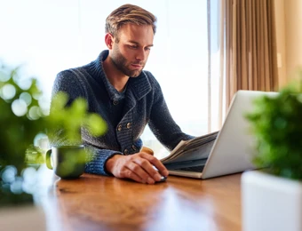 Handsome modern man busy reading the newspaper and checking his mails to catch up on current affairs from the comfort of his home while seated at his dining table.