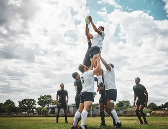 Rugbyspieler heben einen Mitspieler hoch, um den Ball zu fangen; weitere Spieler stehen auf dem Spielfeld.