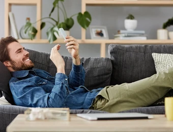Junger Mann liegt auf grauem Sofa, schaut auf Smartphone; Wohnzimmer mit Regal, Pflanzen und Büchern im Hintergrund.