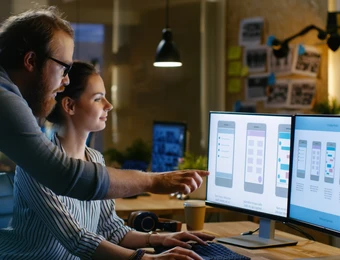 Zwei Personen arbeiten an einem Schreibtisch mit zwei Monitoren, die UI-Entwürfe zeigen; Kaffee und Kopfhörer liegen daneben.