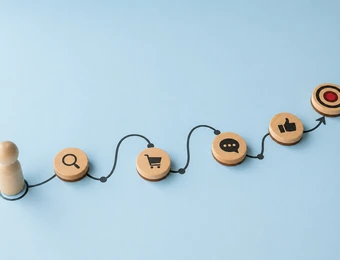 Blauer Hintergrund; zwei Holzfiguren links; Holzplättchen mit Symbolen Lupe, Warenkorb, Chat, Daumen hoch, Ziel über Schnur verbunden.