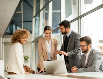 Vier Personen in Business-Outfits stehen um einen Laptop auf einem weißen Tisch in einem modernen Büro.
