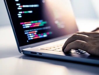 Nahaufnahme eines Laptops; Bildschirm zeigt farbigen Code, eine Hand tippt auf der Tastatur.