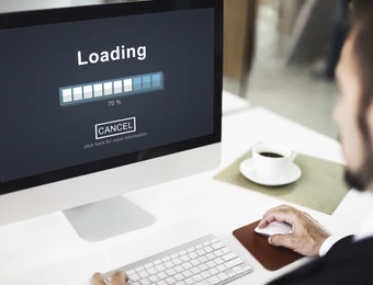 iMac-Bildschirm zeigt Loading mit Fortschrittsbalken ca. 70%; weiße Tastatur, Maus, Kaffeetasse, Person rechts.