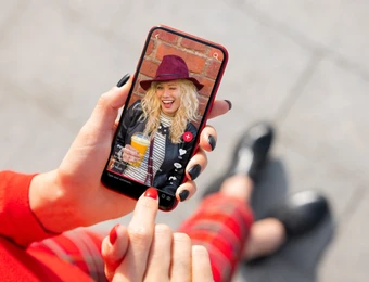 Auf dem Smartphonebildschirm eine blonde Frau mit violettem Hut, Schal und orangefarbenem Getränk vor Backsteinwand.