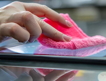 Eine Hand wischt mit einem pinken Mikrofaser-Tuch über eine glatte Bildschirmoberfläche; Spiegelung sichtbar.