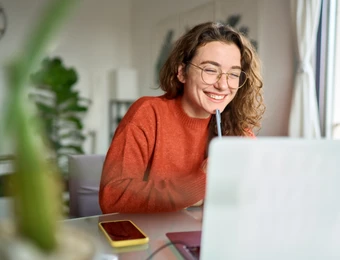 Person mit Brille und orangefarbenem Pullover sitzt am Laptop, lächelt und hält Stift an den Mund.