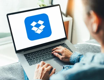 Person tippt auf Laptop; Bildschirm zeigt blaues Dropbox-Logo mit weißem Diamantsymbol.
