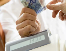 Eine Person hält einen 20-Euro-Schein über einer Registrierkasse; Anzeige und Tastatur sichtbar.