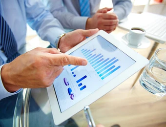 Zwei Personen in Business-Outfit schauen auf ein Tablet mit Balken- und Tortendiagrammen; auf dem Tisch liegen Laptop und Glas Wasser.