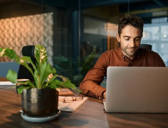 Mann sitzt an Schreibtisch mit Laptop, Notizbuch und Brille; Pflanze im Vordergrund, Bürohintergrund.
