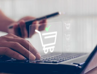 Nahaufnahme von Händen auf Laptop-Tastatur, mit durchsichtigen Einkaufswagen-Symbol und grünem Häkchen.