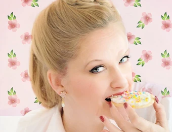 Frau mit blondem Haar in Hochsteckfrisur isst Donut mit bunten Streuseln; rosa Blumenmuster im Hintergrund.