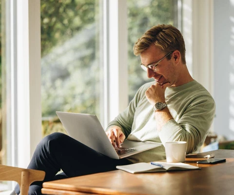 Junger Mann mit Brille sitzt im hellen Raum am Laptop; auf dem Tisch Tasse, Notizbuch und Smartphone.