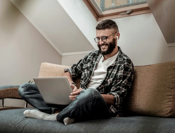 Ein Mann mit Bart und Brille sitzt auf einem Sofa, Laptop auf dem Schoß, trägt kariertes Hemd und Jeans.