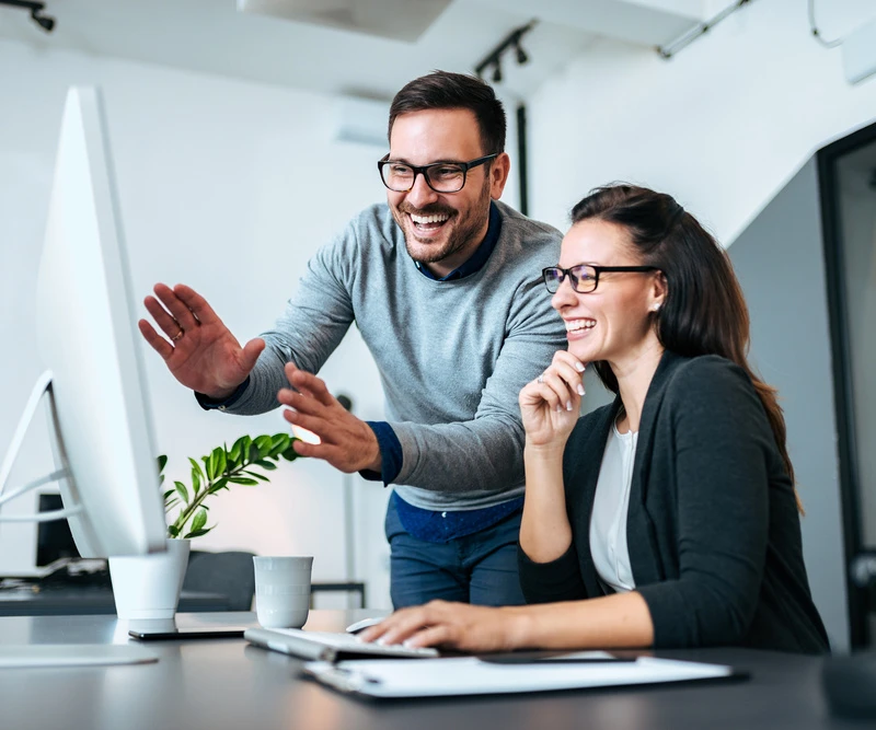 Zwei Personen arbeiten in einem hellen Büro; der Mann steht und gestikuliert, die Frau sitzt am Computer und lächelt.