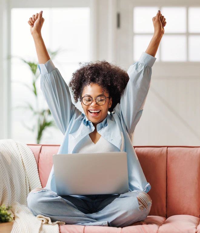 Eine Person sitzt auf einem rosa Sofa, Laptop auf dem Schoß, Arme nach oben gestreckt, lächelnd, Brille, helles Zimmer.