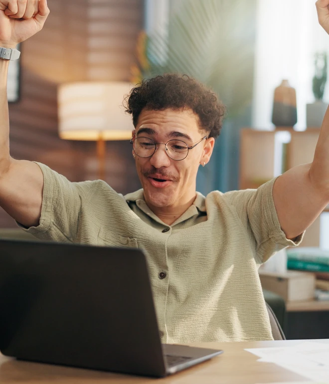 Ein Mann mit Brille sitzt vor Laptop, hebt jubelnd beide Arme hoch; helles Zimmer im Hintergrund.