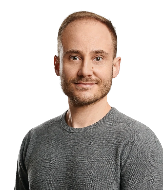 Mann mit grauem Pullover, kurzes braunes Haar, leichter Bart, blickt in die Kamera.