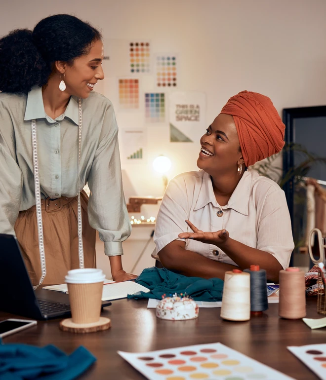 Zwei Frauen in einem Designstudio; eine mit Maßband steht, die andere sitzt mit Turban, umgeben von Stoffen und Nähzubehör.