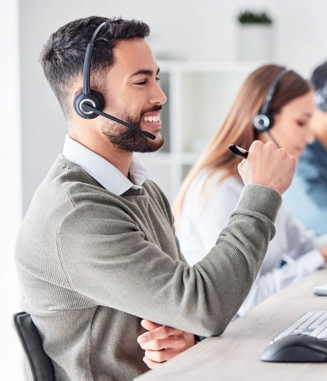 Mann mit Headset und Mikro am Schreibtisch, lächelnd; Hintergrund: weitere Person mit Headset.