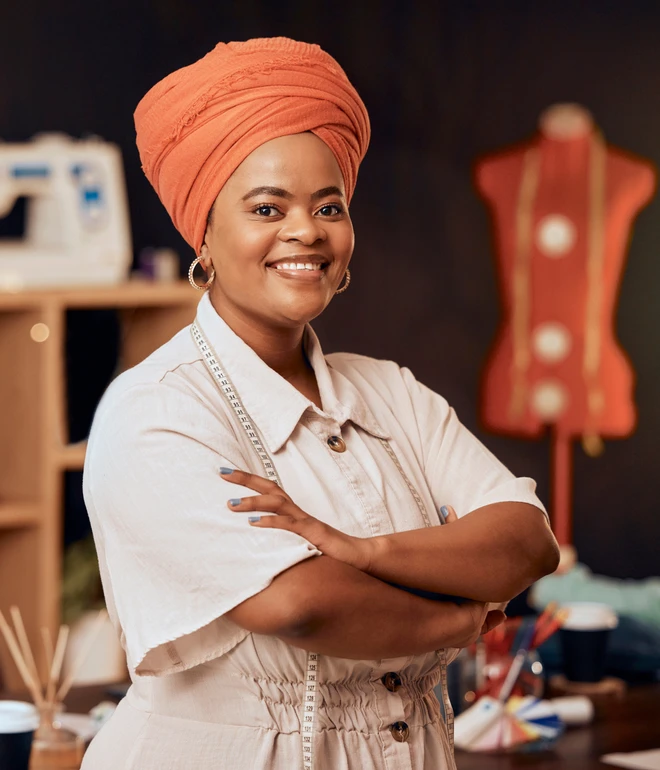 Eine lächelnde Frau mit orangefarbenem Turban, weißem Hemd und Maßband um den Hals; Nähmaschine und Schneiderpuppe.