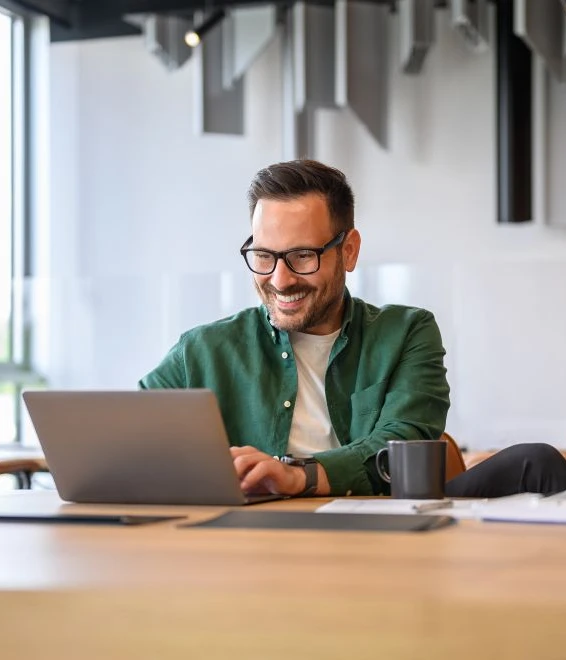 Mann mit Brille in grünem Hemd sitzt am Tisch vor Laptop, Kaffeebecher und Unterlagen; lächelt in hellem Büroraum.