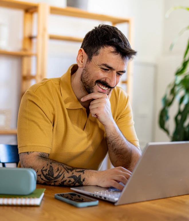 Mann in gelbem Polo sitzt am Holztisch vor Laptop, Kinn gestützt; Notizblock links, grüne Box, Tasse rechts, Regal, Pflanze.
