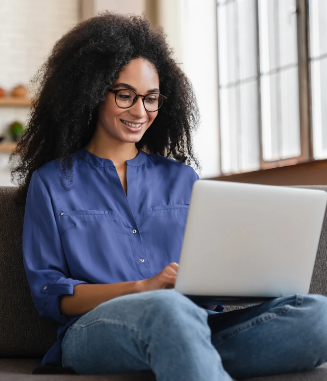 Frau mit lockigem Haar, Brille, blaue Bluse und Jeans sitzt auf einer Couch und nutzt einen Laptop.