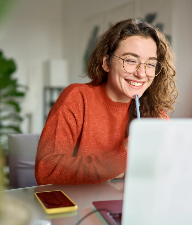 Person mit Brille und orangefarbenem Pullover sitzt am Laptop, lächelt und hält Stift an den Mund.
