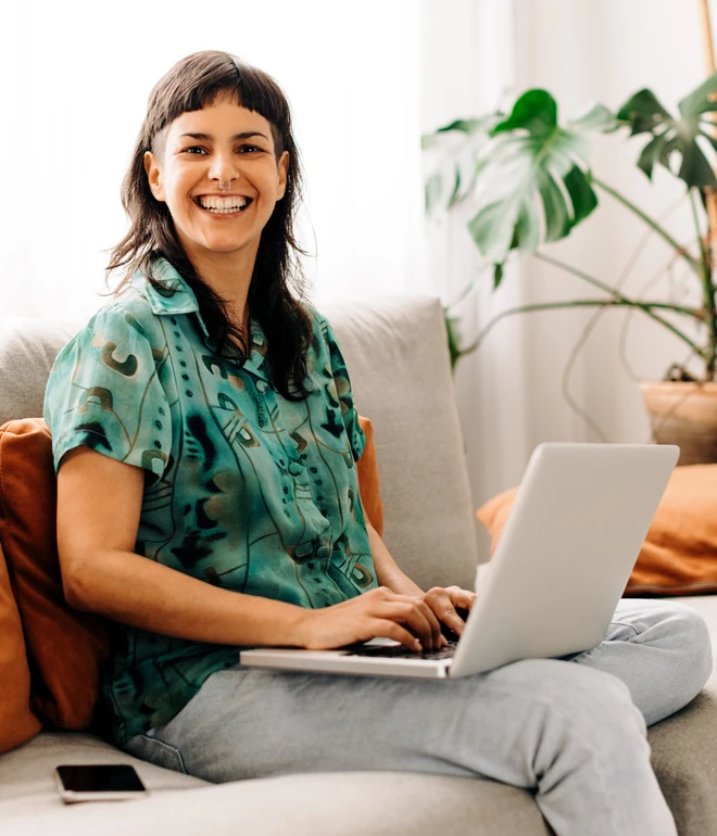 Eine Frau sitzt auf einer hellen Couch mit orangefarbenen Kissen, Laptop auf dem Schoß und Smartphone daneben.