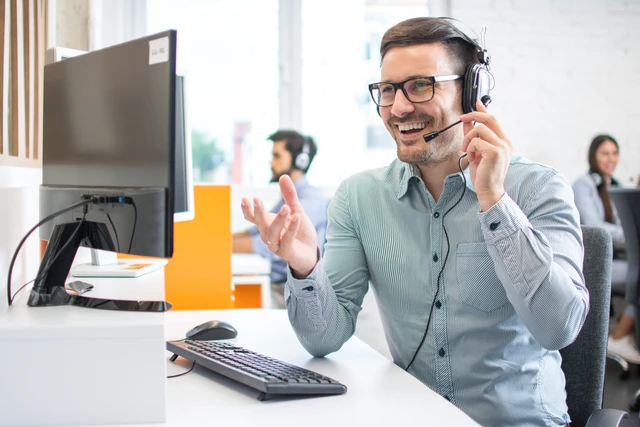 Mann mit Headset sitzt an Schreibtisch vor Monitor, Tastatur und Maus; Büroumgebung mit weiteren Personen im Hintergrund.