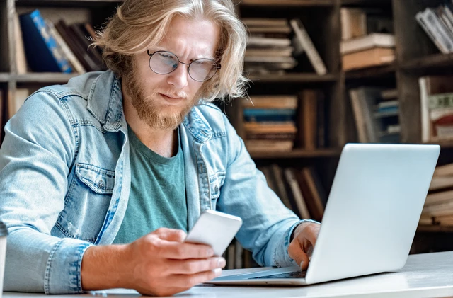 Mann mit Brille und Bart sitzt am Tisch, schaut auf Smartphone, Laptop davor; Bücherregale im Hintergrund.