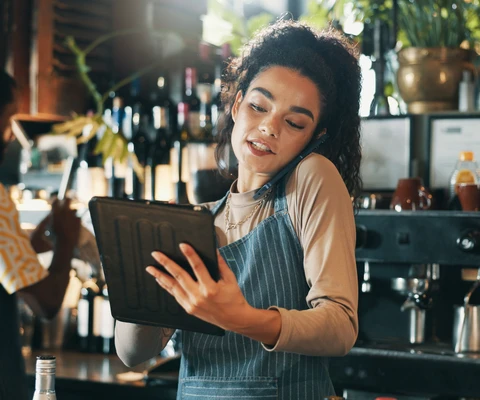 Frau mit blauer Schürze hält Tablet; Bar mit Flaschen im Hintergrund.