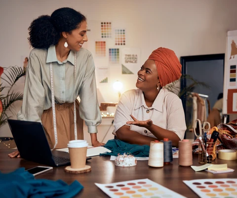 Zwei Frauen in einem Designstudio; eine mit Maßband steht, die andere sitzt mit Turban, umgeben von Stoffen und Nähzubehör.