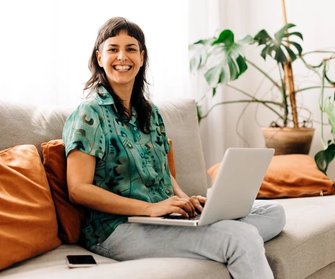 Eine Frau sitzt auf einer hellen Couch mit orangefarbenen Kissen, Laptop auf dem Schoß und Smartphone daneben.