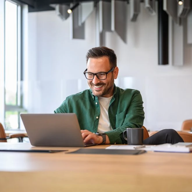 Mann mit Brille in grünem Hemd sitzt am Tisch vor Laptop, Kaffeebecher und Unterlagen; lächelt in hellem Büroraum.