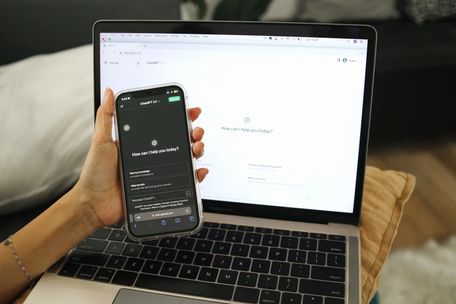 Eine Hand hält ein Smartphone neben einem offenen Laptop; Laptop-Bildschirm zeigt eine Chat-Oberfläche.