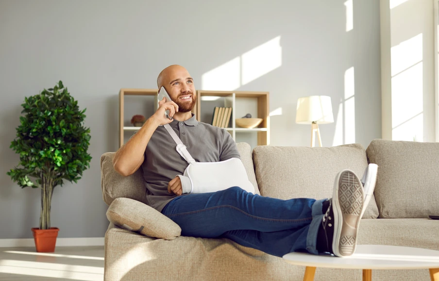 Bärtiger Mann sitzt auf einer Couch, telefoniert, hat Papiere auf dem Schoß; Wohnzimmer mit Pflanze, Regal, Sonnenlicht.