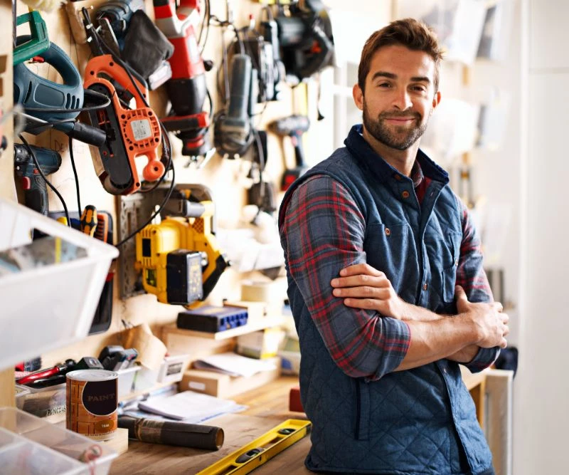Mann mit verschränkten Armen in einer Werkstatt; an der Wand hängen Bohrer, Sägen und Werkzeuge.