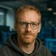 Porträt eines Mannes mit rotem Bart, Brille und Hoodie; neutraler Gesichtsausdruck, unscharfer Hintergrund mit blauen Bildschirmen.