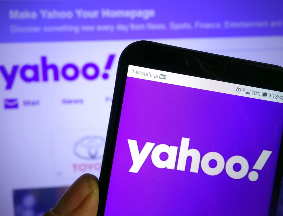 Eine Hand hält ein Smartphone mit lila Bildschirm, der 'yahoo!' zeigt; dahinter ist ein Monitor mit Yahoo-Logo.