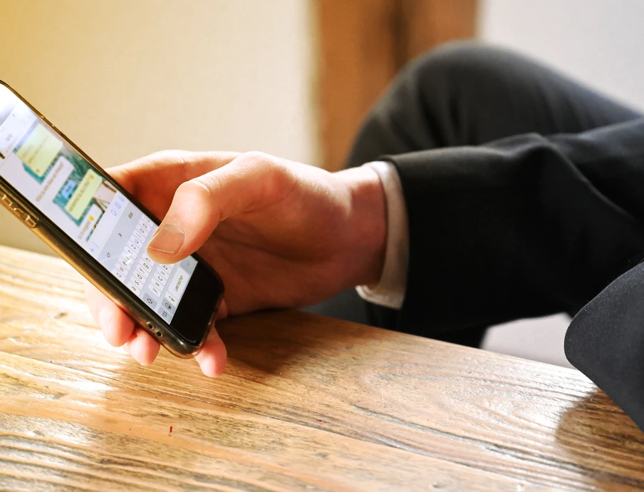 Person im Anzug hält Smartphone über Holztisch; Bildschirm sichtbar.